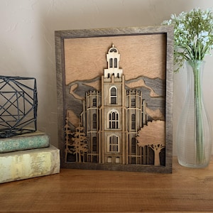 May include: A framed wooden art piece depicting a detailed architectural design of a building with a tall tower. The artwork is layered with various shades of brown and gray, creating a 3D effect. The frame is a dark brown color.