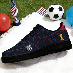 May include: Navy blue and red Nike Air Force 1 trainers with a USA shield logo and star pattern. The shoe features a black sole, red accents, and a gold trophy graphic. A football and balloons are in the background.