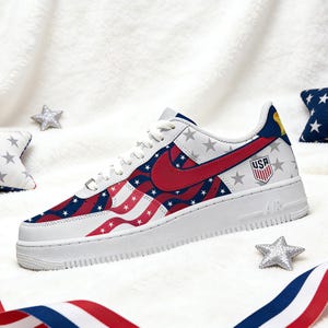 May include: White trainers with a patriotic design featuring red and blue stripes and white stars. The Nike swoosh is red. The heel displays a gold emblem and the word "USA".