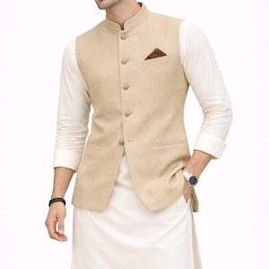 May include: A beige waistcoat with a mandarin collar and a brown pocket square. The waistcoat is worn over a white long-sleeved shirt and white pants. A black bracelet and a silver watch are visible.