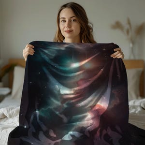 May include: A dark blanket with a vibrant space-themed design. The blanket features a nebula of blues, greens, and reds, with bright star-like highlights. The blanket is held up by a person.