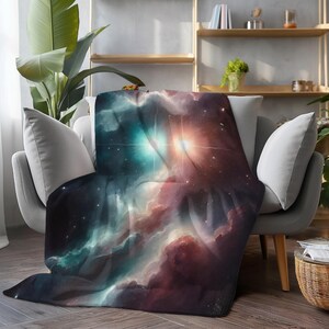 Könnte beinhalten: Eine weiche, flauschige Decke mit einer lebendigen Darstellung eines Weltraumnebels. Das Design zeigt wirbelnde Wolken in Türkis-, Lila- und Orangetönen mit hellen, sternartigen Lichtblitzen. Die Decke ist über einem grauen Sessel drapiert.