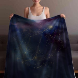 May include: A dark blue blanket with a celestial design featuring constellations and golden stars. The blanket has a soft, plush appearance and is held up by a person. The blanket's design includes shades of purple and blue.