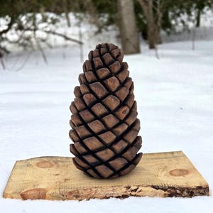 May include: A finely carved wooden pinecone with a detailed diamond pattern, displayed on a light-coloured wooden plank. The background features a snowy landscape and blurred trees. The pinecone is a deep brown colour, creating a natural, rustic aesthetic.