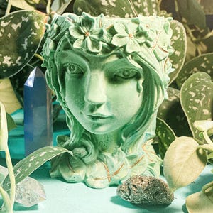 May include: A green ceramic planter shaped like a woman's head with a floral crown. The planter is sitting on a light blue surface with green plants and a crystal.