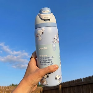 May include: A light blue Owala water bottle featuring Snoopy illustrations. The bottle has a push-button lid and a built-in straw. Text on the bottle reads, "Hi there. I'm leak proof. I'm insulated. And I have a built-in straw."