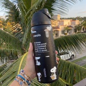 May include: Black Owala water bottle with Snoopy graphics and text. The bottle has a built-in straw and is insulated. The text on the bottle reads "Hi there. I'm leak proof. I'm insulated. And I have a built-in straw."