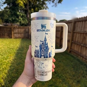 May include: A cream-colored Stanley Quencher H2.0 tumbler with a white handle and lid. The tumbler features a blue castle design, fireworks, and the Stanley logo. The text "The Flowstate Quencher H2.0 Tumbler" is printed on the bottom.