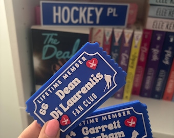 Op de afbeelding: Twee blauwe fanclubtickets met witte tekst en rode hart- en hockeystickafbeeldingen. Op één ticket staat "Dean Di Laurentis Fan Club" en op de andere "Garrett Graham Fan Club". Op de achtergrond is een blauw "HOCKEY PL"-bord zichtbaar.