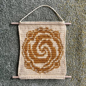 May include: A beige and gold crochet wall hanging with a spiral design. The handmade piece features a geometric pattern in shades of beige and gold, with a wooden dowel at the top and bottom for hanging. The wall hanging is approximately 10 inches wide and 12 inches long.