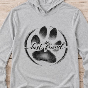 May include: A light gray long-sleeve hooded shirt featuring a black circular graphic on the chest. The graphic contains a textured paw print design with the words best friend written in a cursive script across the center of the paw print.