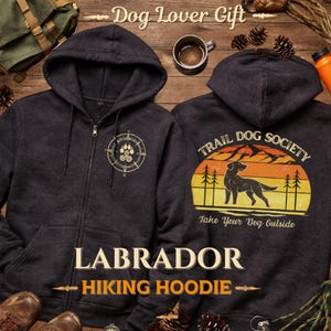 May include: Dark gray Labrador hiking hoodie with a compass and paw print design on the front. The back features a sunset graphic with a dog silhouette and the text "Trail Dog Society". The words "Dog Lover Gift" are at the top.