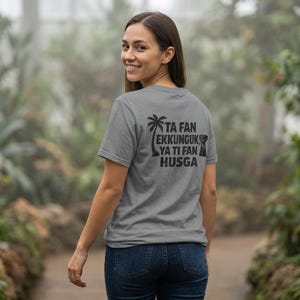 Könnte beinhalten: Ein graues T-Shirt mit schwarzem Text und einer Palmen-Grafik. Der Text lautet "TA FAN EKKUNGUK YA TI FAN HUSGA". Das T-Shirt wird von einer Person in blauen Jeans getragen, die in die Kamera lächelt.