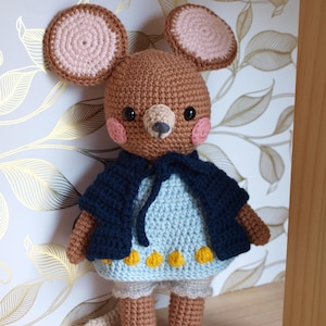 May include: A handmade crochet mouse doll with large, round ears. The mouse wears a blue dress with yellow accents and a navy blue cape. The doll has a light brown body, pink cheeks, and a small tail.