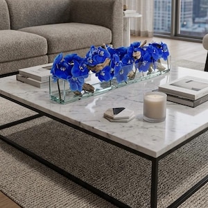 May include: A modern living room with a marble coffee table featuring a rectangular glass vase filled with vibrant blue orchids and driftwood. A lit candle and books are also on the table. The table has a black metal frame.