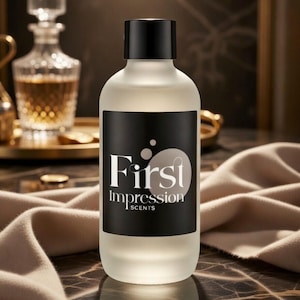 May include: A frosted glass perfume bottle with a black cap and a black label. The label reads "First Impression SCENTS" in white and silver lettering. The bottle is set against a blurred background with a decanter and a tray.