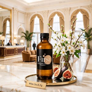 May include: An amber glass bottle with a black cap and a label reading First Impression Scents. Next to it is a clear glass vase containing white flowers and green stems, accompanied by sliced figs on a gold tray with a White Tea & Fig label.