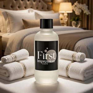 May include: A frosted glass bottle with a black cap and a black label that reads "First Impression SCENTS" is centered between two rolled white towels. The background features a bed with white linens and a lamp.