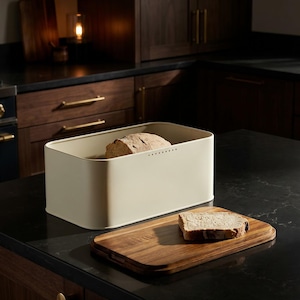 Peut inclure: Un coffre à pain crème contenant un pain, à côté d'une planche à découper en bois avec une tranche de pain sur un comptoir noir. Le coffre à pain est rectangulaire avec des coins arrondis et une rangée de petits trous sur le dessus.