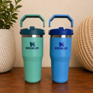 May include: Two Stanley insulated tumblers, one teal and one blue, with matching handles and lids. Each tumbler features the Stanley logo and a winged creature emblem. The tumblers are designed for beverages and are shown on a wooden surface.