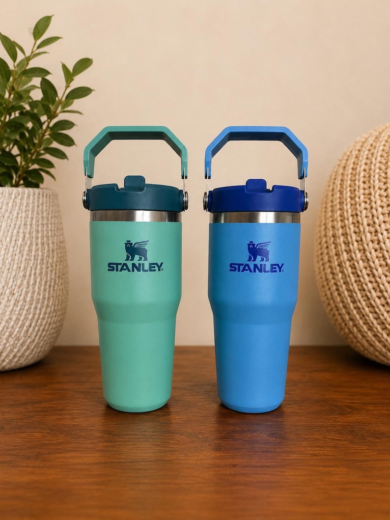 May include: Two Stanley insulated tumblers, one teal and one blue, with matching handles and lids. Each tumbler features the Stanley logo and a winged creature emblem. The tumblers are designed for beverages and are shown on a wooden surface.