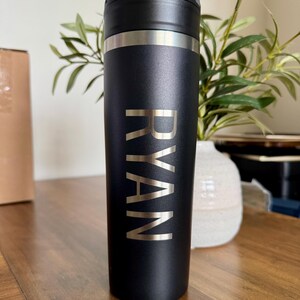 K&ouml;nnte beinhalten: Ein zylindrischer Trinkbecher aus mattschwarzem Edelstahl mit dem vertikal eingravierten Namen RYAN in einer klaren serifenlosen Schrift. Das Gef&auml;&szlig; verf&uuml;gt &uuml;ber einen schwarzen Deckel mit integriertem Griff und ein silberfarbenes Metallband am oberen Rand.