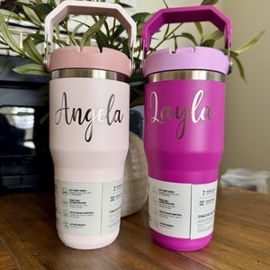 K&ouml;nnte beinhalten: Zwei isolierte Trinkbecher mit Griffen, einer in Hellrosa mit dem Namen Angela und einer in Magenta mit dem Namen Layla. Beide verf&uuml;gen &uuml;ber einen silbernen Metallrand, einen Tragegriff und ein wei&szlig;es Produktinformationsetikett.