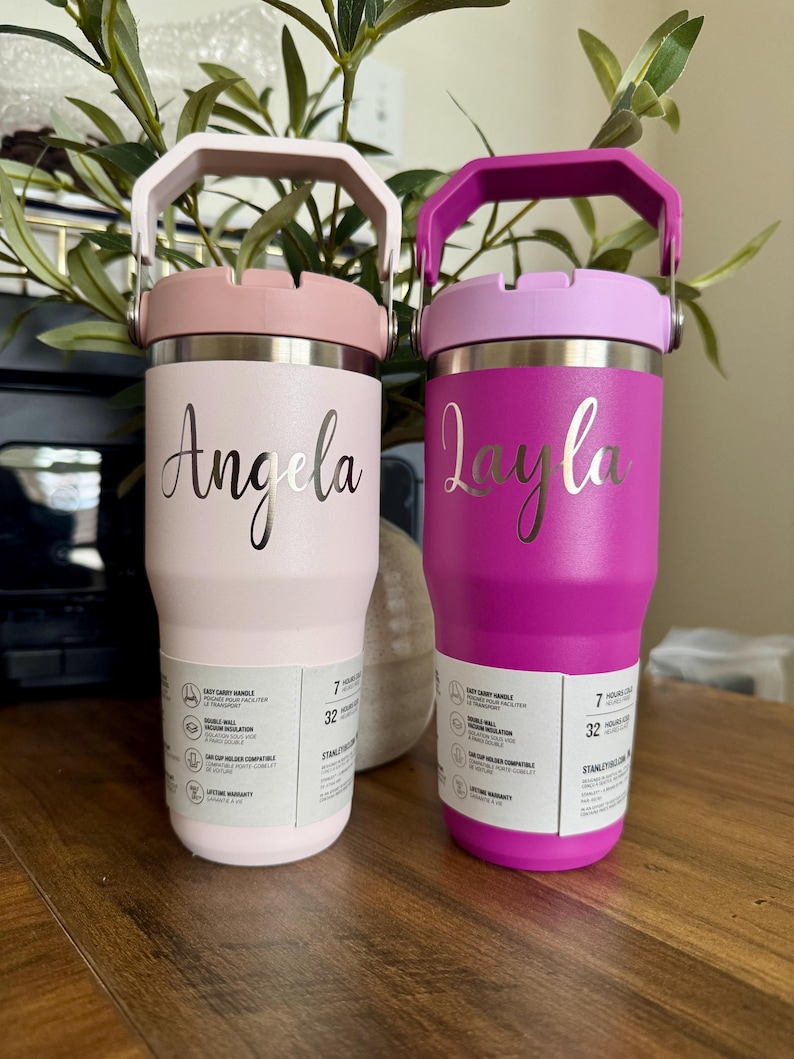K&ouml;nnte beinhalten: Zwei isolierte Trinkbecher mit Griffen, einer in Hellrosa mit dem Namen Angela und einer in Magenta mit dem Namen Layla. Beide verf&uuml;gen &uuml;ber einen silbernen Metallrand, einen Tragegriff und ein wei&szlig;es Produktinformationsetikett.