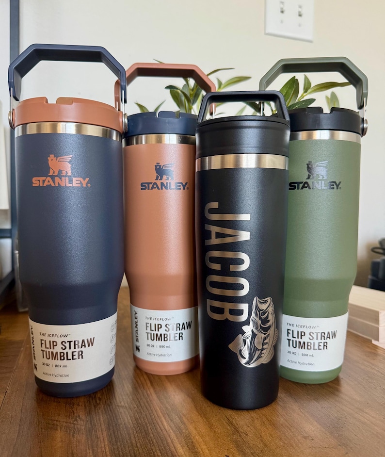 K&ouml;nnte beinhalten: Vier Stanley IceFlow Flip Straw Becher in Marineblau, Pfirsich, Schwarz und Olivgr&uuml;n. Jeder 887 ml Becher hat einen Griff, einen Deckel und ein Etikett. Der schwarze Becher ist mit dem Namen JACOB und einer Fischgrafik personalisiert.