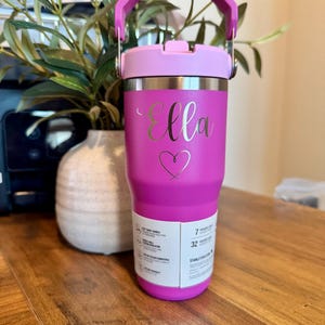 K&ouml;nnte beinhalten: Ein leuchtend pinkfarbener Isolierbecher mit passendem Griff und Deckel. Der Name Ella ist in Schreibschrift &uuml;ber einem Herzumriss eingraviert. Ein wei&szlig;es Produktetikett an der unteren Vorderseite zeigt Merkmale wie 7 Stunden kalt und 32 Stunden mit Eis.