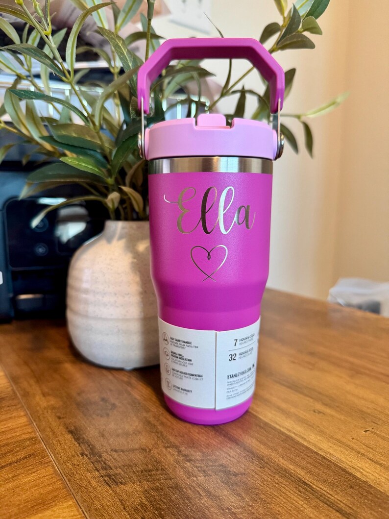 K&ouml;nnte beinhalten: Ein leuchtend pinkfarbener Isolierbecher mit passendem Griff und Deckel. Der Name Ella ist in Schreibschrift &uuml;ber einem Herzumriss eingraviert. Ein wei&szlig;es Produktetikett an der unteren Vorderseite zeigt Merkmale wie 7 Stunden kalt und 32 Stunden mit Eis.