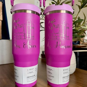 K&ouml;nnte beinhalten: Zwei leuchtend pinke Edelstahlbecher mit violetten Deckeln und Griffen. Jeder zeigt ein lasergraviertes Design aus B&uuml;chern, einem Apfel und dem Text "ABC 123". Einer ist mit "Mrs. Arxer" und der andere mit "Ms. Passante" in Schreibschrift personalisiert.