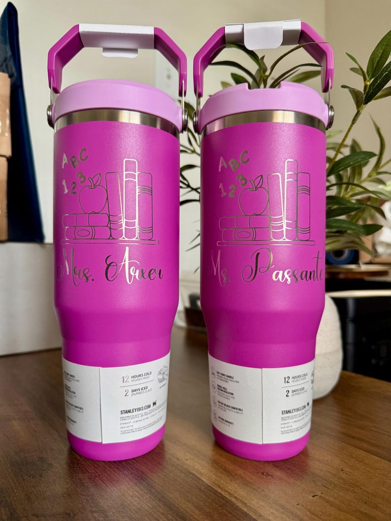 K&ouml;nnte beinhalten: Zwei leuchtend pinke Edelstahlbecher mit violetten Deckeln und Griffen. Jeder zeigt ein lasergraviertes Design aus B&uuml;chern, einem Apfel und dem Text "ABC 123". Einer ist mit "Mrs. Arxer" und der andere mit "Ms. Passante" in Schreibschrift personalisiert.