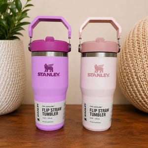May include: Two Stanley Iceflow Flip Straw Tumblers, one in light purple and the other in light pink. Each tumbler has a matching handle and lid, with the Stanley logo and product information printed on the front. Each tumbler holds 14 oz / 410 ml.