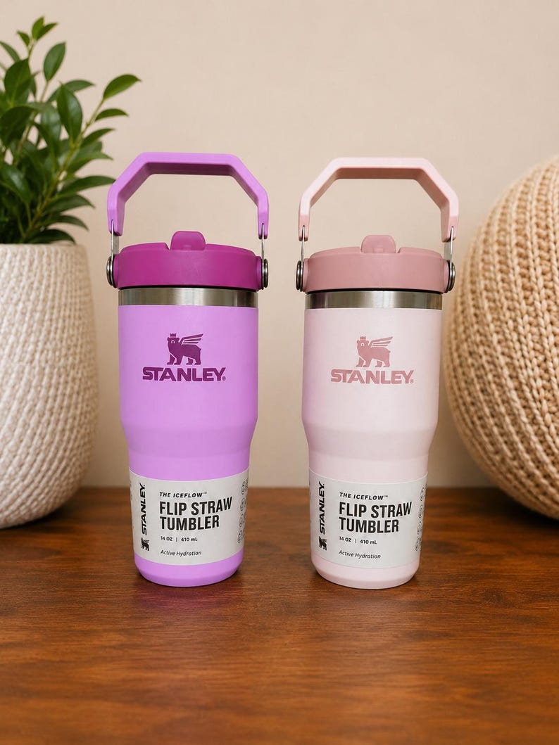 May include: Two Stanley Iceflow Flip Straw Tumblers, one in light purple and the other in light pink. Each tumbler has a matching handle and lid, with the Stanley logo and product information printed on the front. Each tumbler holds 14 oz / 410 ml.