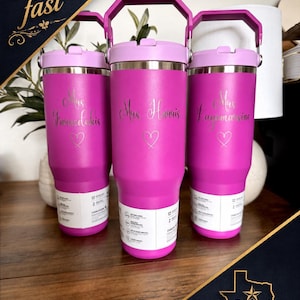 K&ouml;nnte beinhalten: Drei leuchtend pinke Edelstahlbecher mit violetten Deckeln und Griffen. Jeder Becher ist mit einem in Schreibschrift gravierten Namen &uuml;ber einem kleinen Herzumriss versehen. Ein wei&szlig;es Produktetikett ist um den unteren Teil gewickelt.