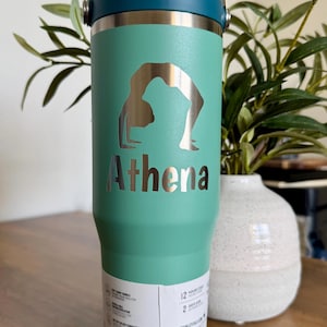 K&ouml;nnte beinhalten: Ein t&uuml;rkisfarbener Isolierbecher mit dunkelblauem Deckel und Griff. Auf der Vorderseite befindet sich eine silberfarbene Silhouette einer Person in einer Yoga-Br&uuml;ckenpose &uuml;ber dem Namen Athena in fetter, dunkler Schrift. Ein wei&szlig;es Etikett ist angebracht.