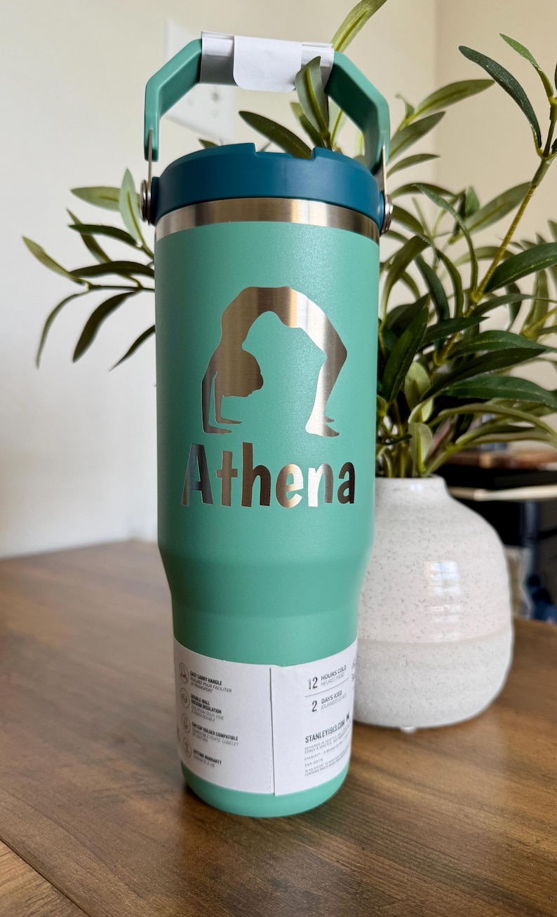 K&ouml;nnte beinhalten: Ein t&uuml;rkisfarbener Isolierbecher mit dunkelblauem Deckel und Griff. Auf der Vorderseite befindet sich eine silberfarbene Silhouette einer Person in einer Yoga-Br&uuml;ckenpose &uuml;ber dem Namen Athena in fetter, dunkler Schrift. Ein wei&szlig;es Etikett ist angebracht.