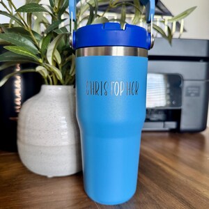 May include: A light blue insulated tumbler with a dark blue lid and handle. The tumbler has the name "CHRISTOPHER" etched on the front. The tumbler is designed to keep beverages cold or hot. The tumbler is approximately 7 inches tall.