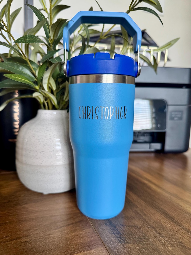 May include: A light blue insulated tumbler with a dark blue lid and handle. The tumbler has the name "CHRISTOPHER" etched on the front. The tumbler is designed to keep beverages cold or hot. The tumbler is approximately 7 inches tall.