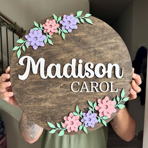 May include: A circular dark brown stained wooden sign featuring the white 3D lettering Madison and CAROL. The sign is decorated with pastel pink and light purple flowers along with light green leaves positioned at the top left and bottom right.