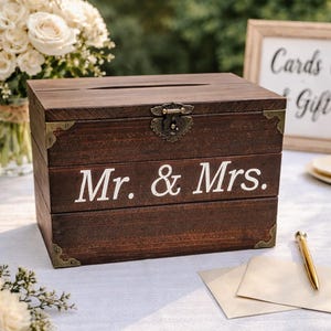 以下が含まれることがあります： 「Mr. & Mrs.」の白い文字が入った、濃い茶色の木製カードボックス。ボックスの上部にはスロットがあり、装飾的な留め金が付いています。コーナーブラケットと白い花のブーケも表示されています。