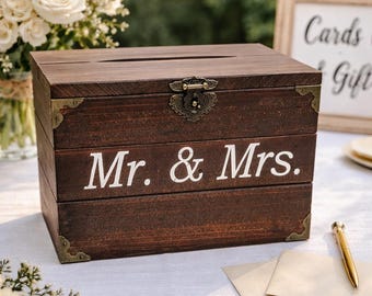 Peut inclure: Un coffret à cartes en bois brun foncé avec les mots "Mr. & Mrs." en lettres blanches. La boîte a une fente sur le dessus et un fermoir décoratif. Des coins et un bouquet de fleurs blanches sont également visibles.
