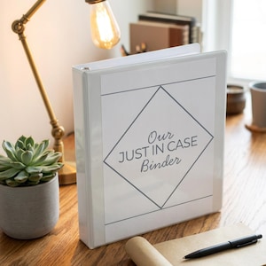 May include: A white binder with the text "Our JUST IN CASE Binder" in a diamond design. The binder is on a wooden desk with a succulent plant, a lamp, and a pen.