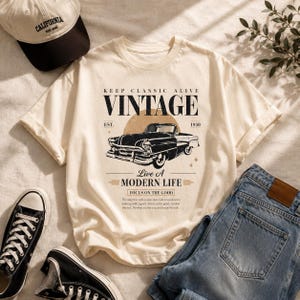 Puede incluir: Una camiseta de color crema con un gráfico negro de un coche antiguo, el texto KEEP CLASSIC ALIVE VINTAGE EST 1950, Live A MODERN LIFE y FOCUS ON THE GOOD, junto con un párrafo de texto pequeño debajo de la ilustración del coche.