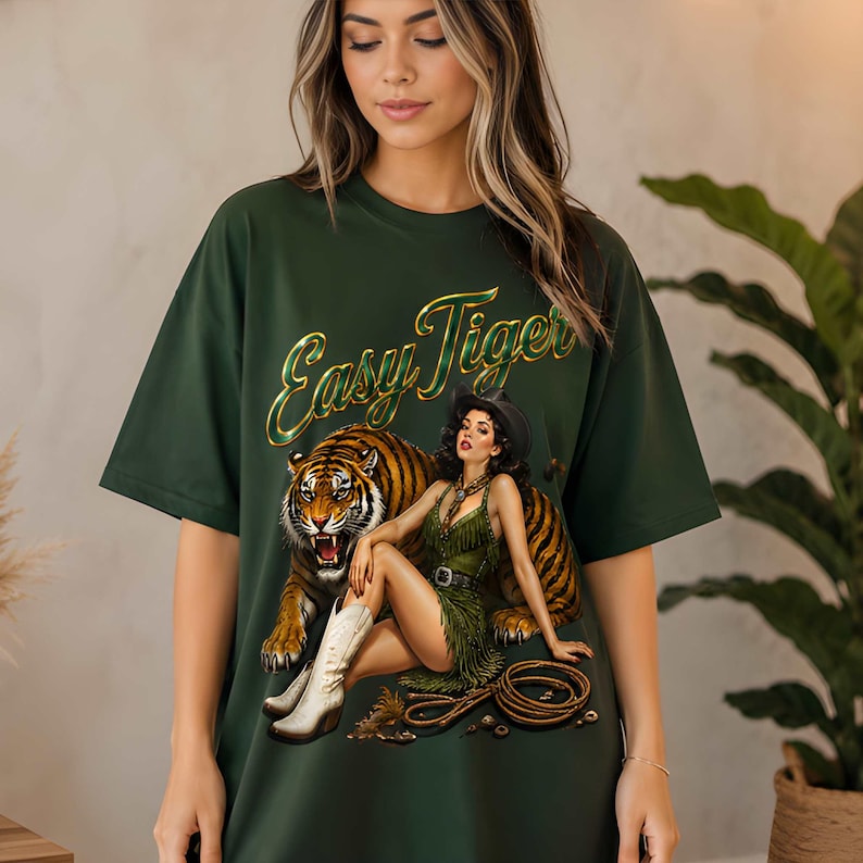 May include: Dark green t-shirt featuring a graphic of a tiger and a woman in a cowgirl outfit. The text "Easy Tiger" is in gold script. The woman is wearing a green fringed dress, a black hat, and white boots.