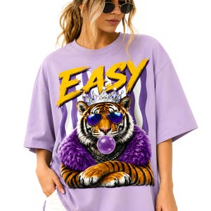 May include: A lilac-colored t-shirt with a graphic of a tiger wearing sunglasses, a crown, and a purple fur collar. The word "EASY" is printed in yellow above the tiger. The shirt has short sleeves and a relaxed fit.