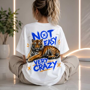 May include: White t-shirt with a tiger graphic and the text "NOT EASY VERY CRAZY" in blue and orange. The tiger is orange and black. The shirt is a casual style.
