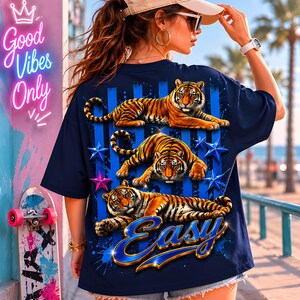 May include: Navy blue t-shirt featuring three orange tigers with black stripes, blue stars, and the word "Easy" in a blue and gold font. The shirt also has blue vertical stripes and a splash effect.