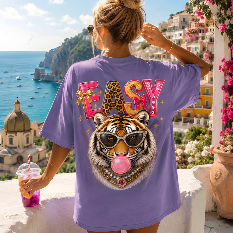 May include: A lilac-colored t-shirt with the word "EASY" in pink and leopard print, featuring a tiger wearing sunglasses and blowing a bubblegum bubble. The tiger has a gold chain necklace with a pink jewel.